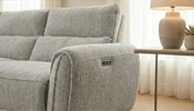 A close-up of a light gray upholstered reclining sofa in a bright living room, showing the side control panel on the armrest and a matching side table with a lamp beside it.