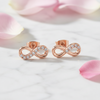 A pair of rose gold infinity loop stud earrings set with small clear crystals, displayed on a white marble surface with soft pink flower petals.